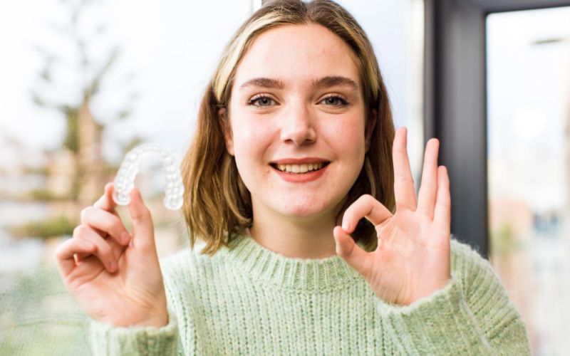 a woman holdf up an invisalign aligner in one hand and make an OK sign with her other hand