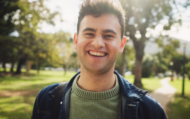 A smiling man stands in a park, surrounded by greenery and trees, enjoying a sunny day.