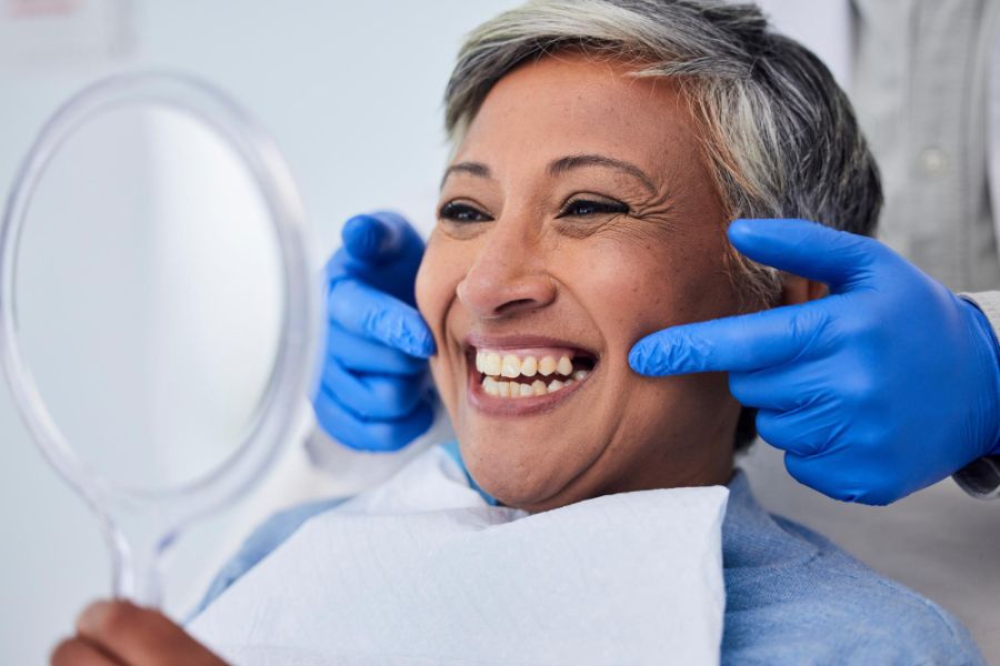 mature woman with grey hair sitting in a dentist chair, she is looking into a mirror and smilining , the dentist is behinfd her wearing blue gloves and pointing to her mouth