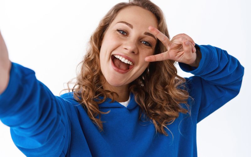 A woman wearing a blue sweatshirt holds up her right hand in a peace sign, smiling at the camera.